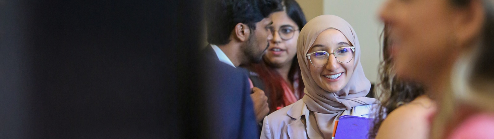 A smiling student in a light pink hijab and glasses holds a purple notebook while talking with other students during a networking event at the University of Florida Warrington College of Business.