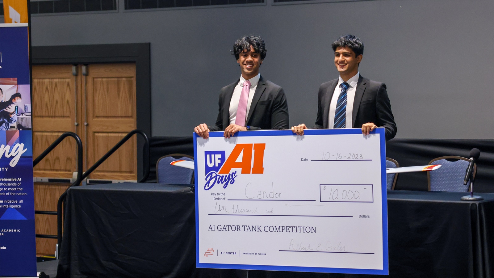 Two young men in suits stand on stage smiling and holding a large ceremonial check for $10,000, awarded for winning the AI Gator Tank Competition at the University of Florida.