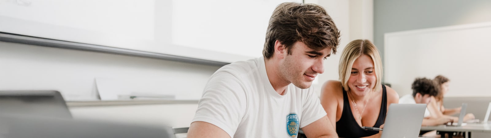 A male student in a white t-shirt and a female student in a black top sit side by side in a classroom, smiling and collaborating as they look at a laptop together.