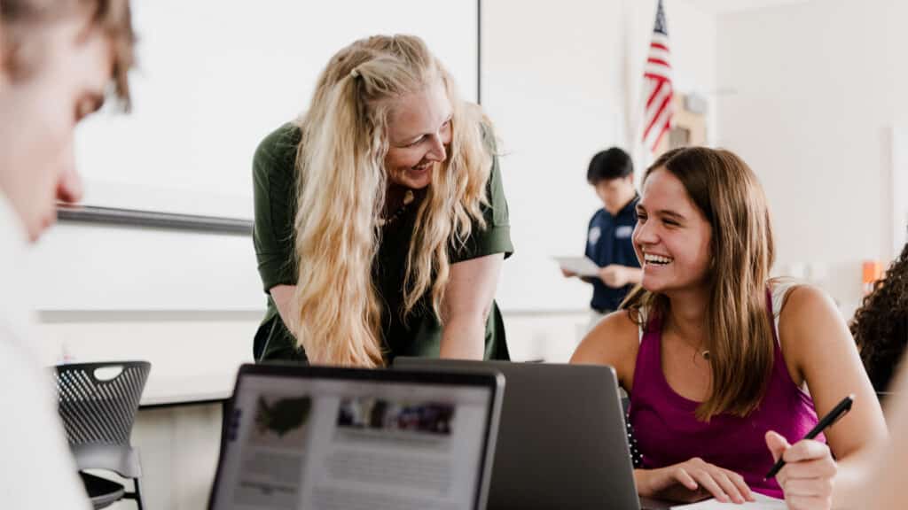A smiling female student in a magenta top sits at a classroom desk with her laptop open, laughing as she talks with a professor who leans in supportively.