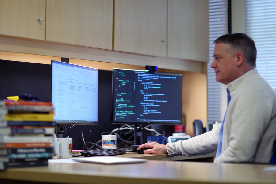 A man in a light sweater sits at an office desk, working on dual monitors—one displaying code and the other email—surrounded by books, mugs, and office supplies.