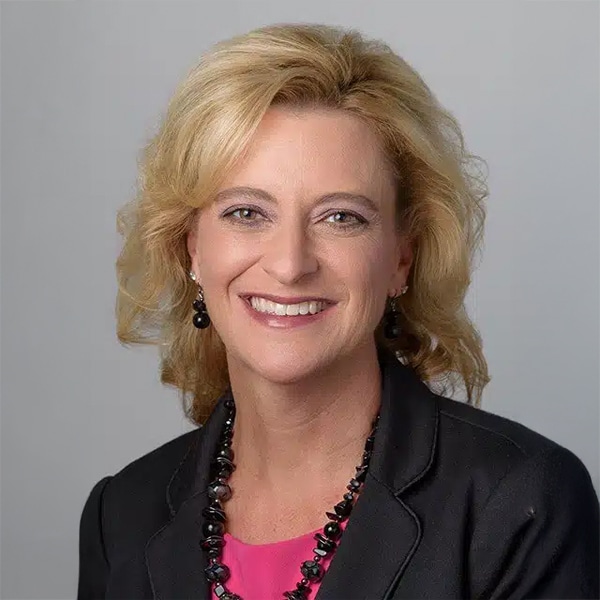 Headshot of Wanda Riley, a smiling woman with shoulder-length blonde hair, wearing a black blazer, pink top, and black beaded necklace, against a neutral gray background.