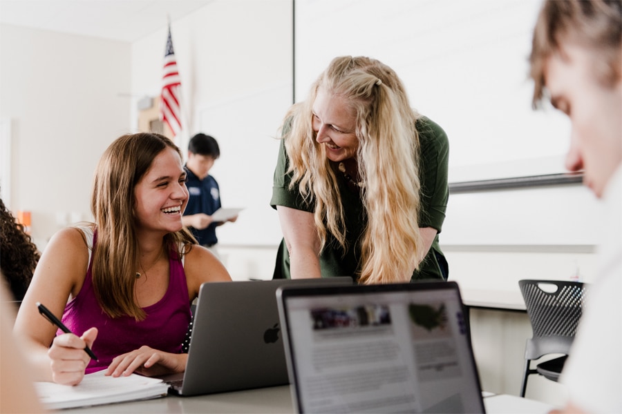 A student laughs while writing notes, engaging with a smiling instructor leaning over her desk in a bright classroom setting.