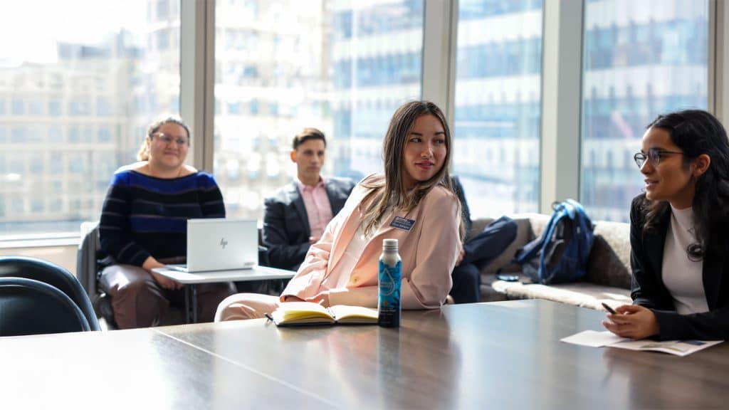 A group of young professionals sit and chat in a bright conference room with tall windows and city views. One woman in a light pink suit is engaged in conversation, holding a notebook.