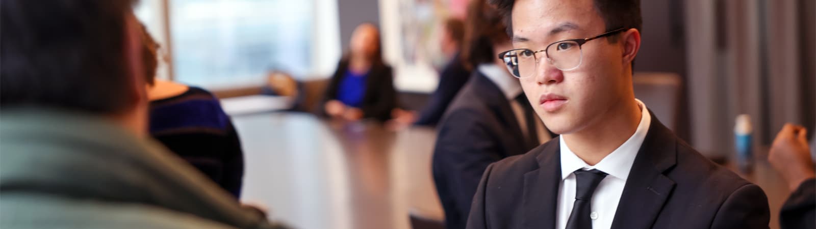 A focused young professional in glasses and a dark suit listens attentively during a business meeting or financial discussion in a modern conference room.