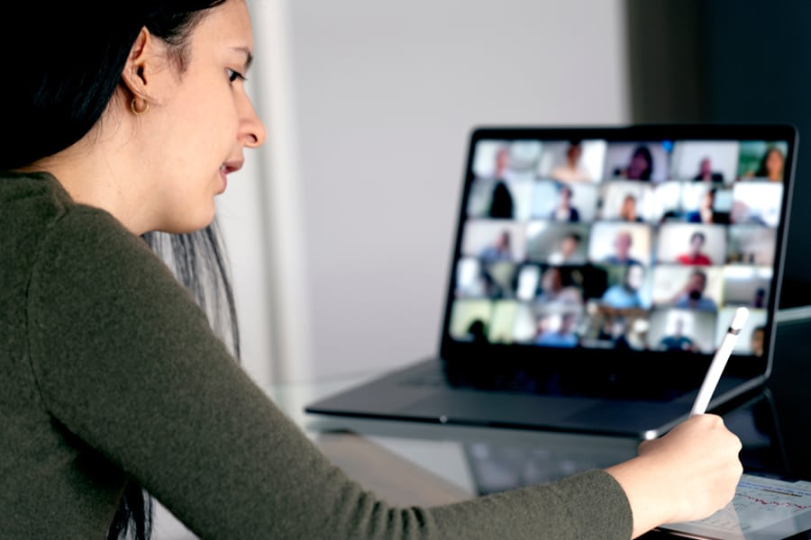 A student participates in an online class discussion, writing notes while viewing a large video conference grid of classmates on a laptop screen.