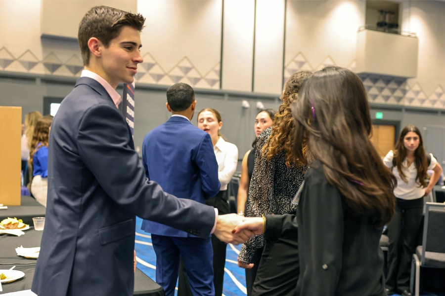A young man in a navy suit shakes hands with a young woman in a black blazer in a lively conference room filled with people networking and talking.