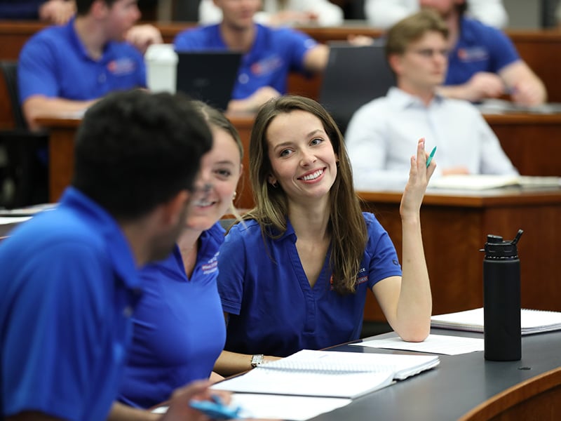 Two young women and a young man seated in a classroom talking to each other with notebooks in front of them and other students in the background.
