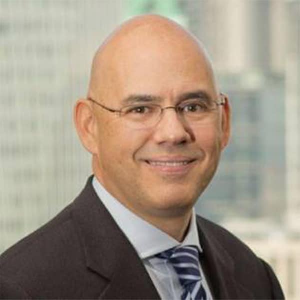 Professional headshot of Alex Abreu, a man wearing glasses, a dark suit jacket, white shirt, and striped tie, smiling with a cityscape background.