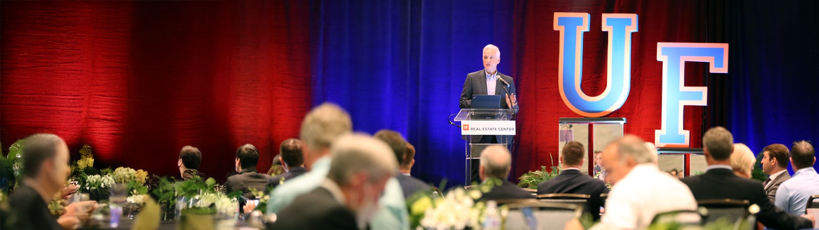 A distinguished speaker stands at a podium labeled "UF Real Estate Center" addressing a seated audience at a formal event. Behind him, large letters "UF" are prominently displayed against a backdrop of red and blue lighting.