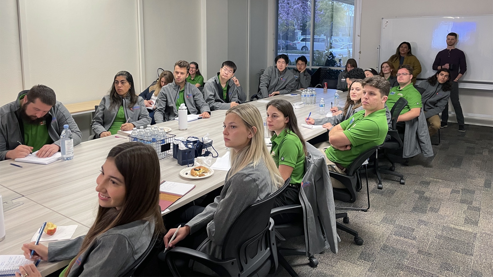 A large group of students and faculty members sit attentively around a rectangular conference table, some taking notes, others listening. Many wear green polo shirts and gray jackets. A whiteboard and large windows are visible in the background.