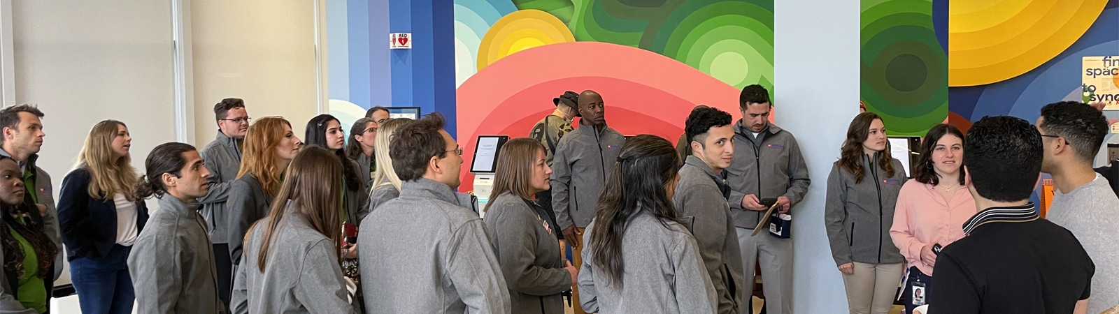 A group of entrepreneurship students and faculty, wearing matching gray jackets, attentively listen during a guided tour in a brightly colored, modern indoor space with a large mural.