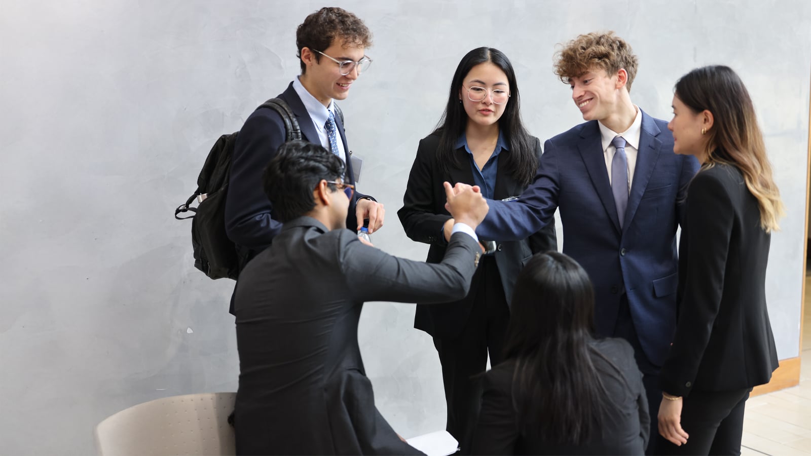 A group of professionally dressed business students gathers indoors around one seated peer. Two students engage in a friendly handshake while others look on and smile.