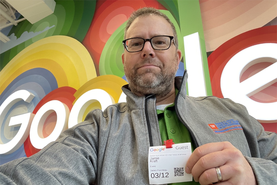 A man wearing glasses and a gray jacket with a University of Florida Warrington College of Business Entrepreneurship & Innovation Center logo poses for a selfie inside the Google office. He holds a visitor badge with the name Jamie Kraft. The colorful Google logo is visible in the background.