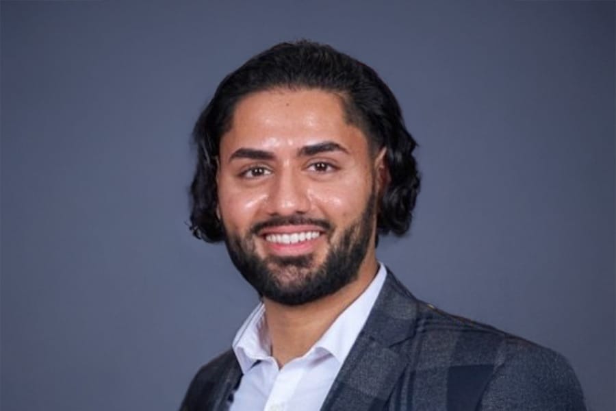 Manpreet Suri, a young man with dark hair and a neatly trimmed beard, smiles confidently while wearing a checkered blazer and white shirt against a plain gray background.