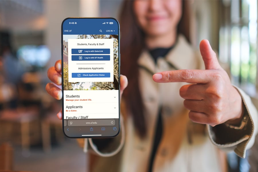 Close-up of a smiling person holding a smartphone displaying the ONE.UF login page, with the screen showing options for students, faculty, staff, and admissions applicants.