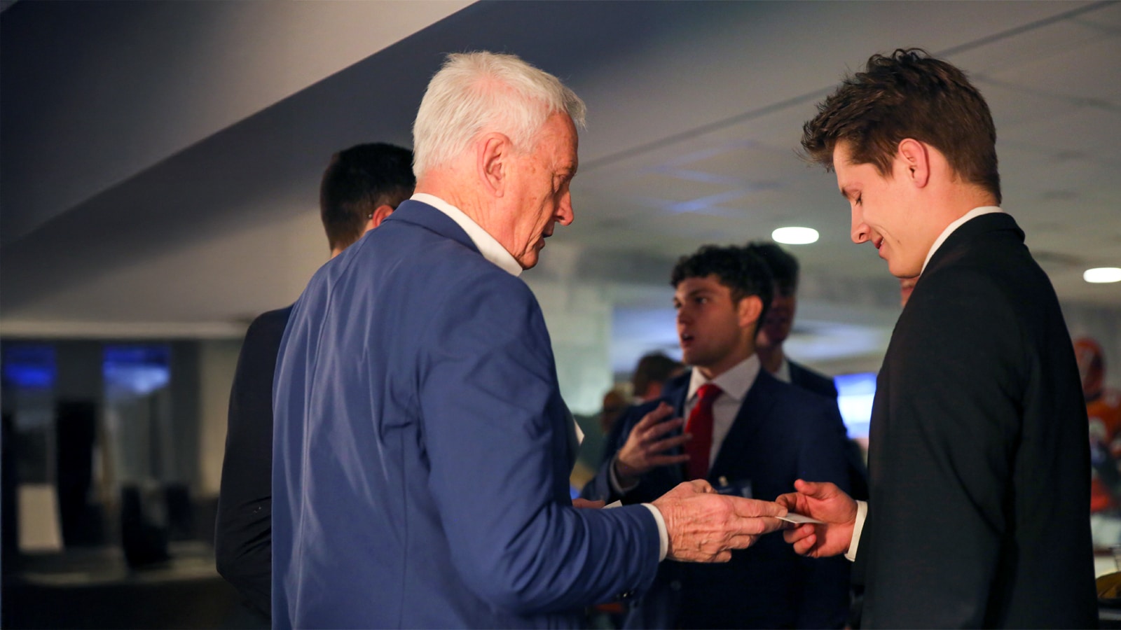 An older male alumnus in a blue blazer exchanges a business card with a younger male student dressed in a black suit, surrounded by other students networking in a professional event setting.