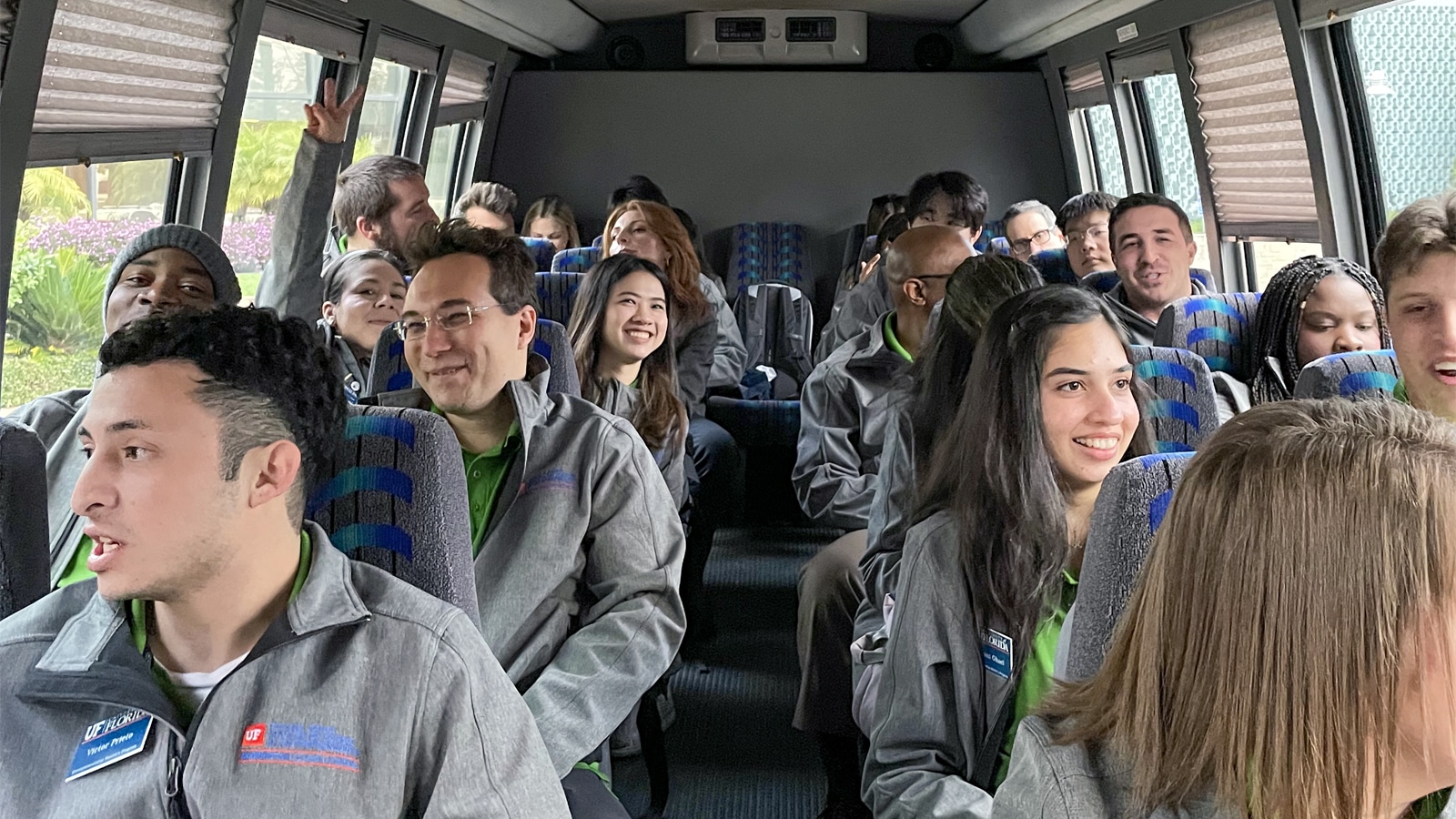 A diverse group of entrepreneurship students and faculty, dressed in matching gray jackets with green shirts underneath, seated inside a bus, smiling and engaging with one another as they travel.