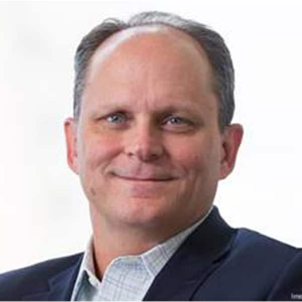 Headshot of Paul Ellis, a middle-aged man with light skin, short light brown hair, wearing a dark blazer over a light checkered shirt, smiling slightly against a plain white background.