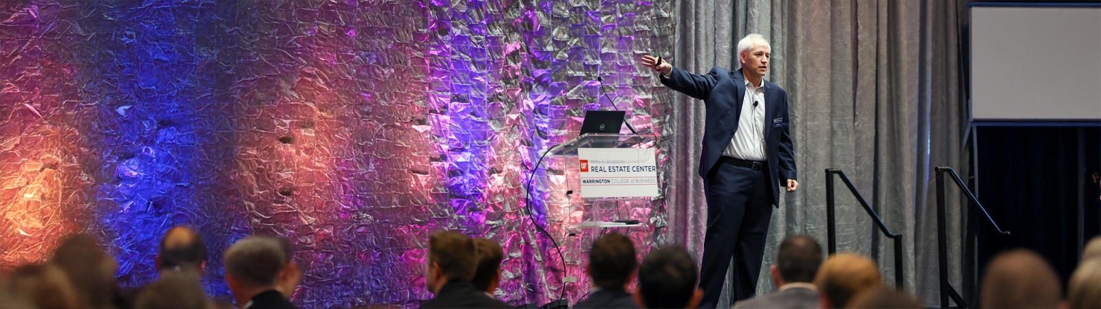 A speaker addresses an engaged audience at a Bergstrom Real Estate Center event, with vibrant purple and orange lighting illuminating the textured backdrop behind him.