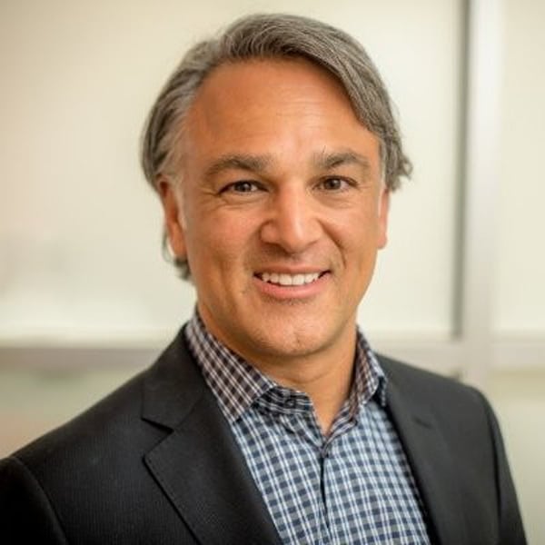 Portrait of Renato Scaff, smiling, with gray hair, wearing a dark blazer over a blue checkered shirt, in front of a softly blurred office background.
