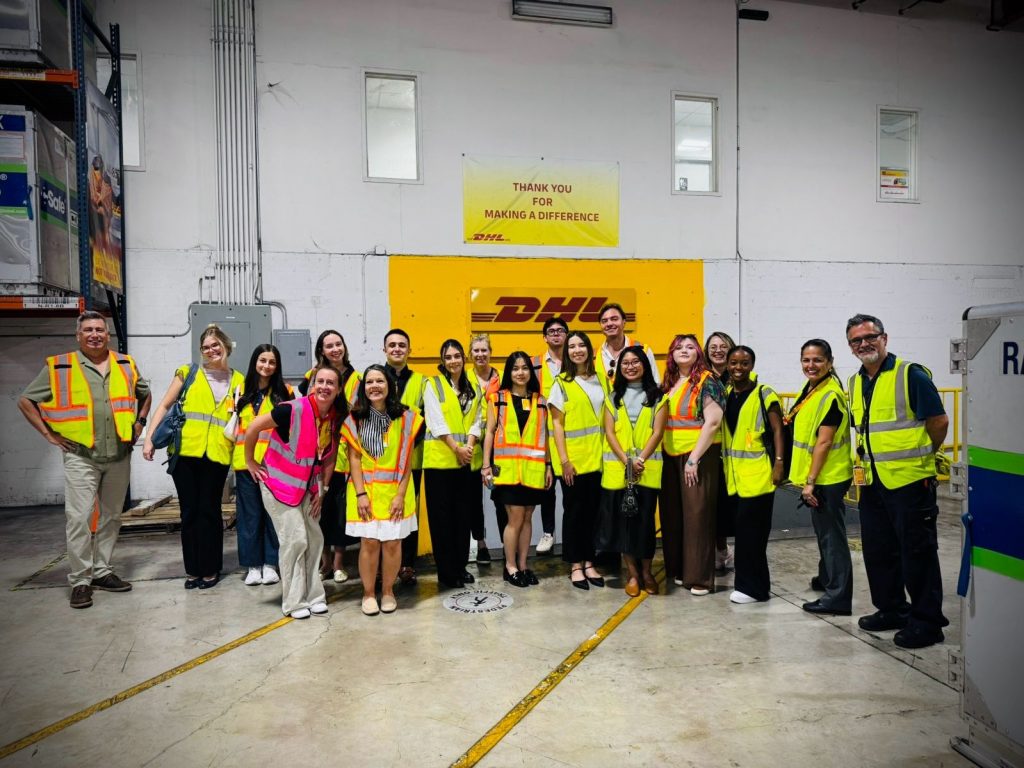 A group of about 20 students wearing yellow safety vests in a DHL warehouse. 