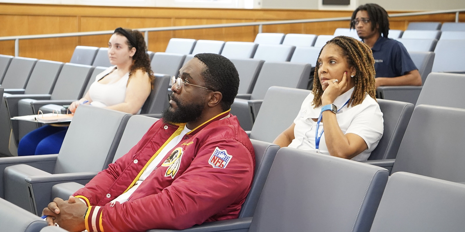 Several individuals seated in a lecture hall, listening attentively, including a man in a red NFL jacket and a woman in a white shirt resting her chin on her hand.
