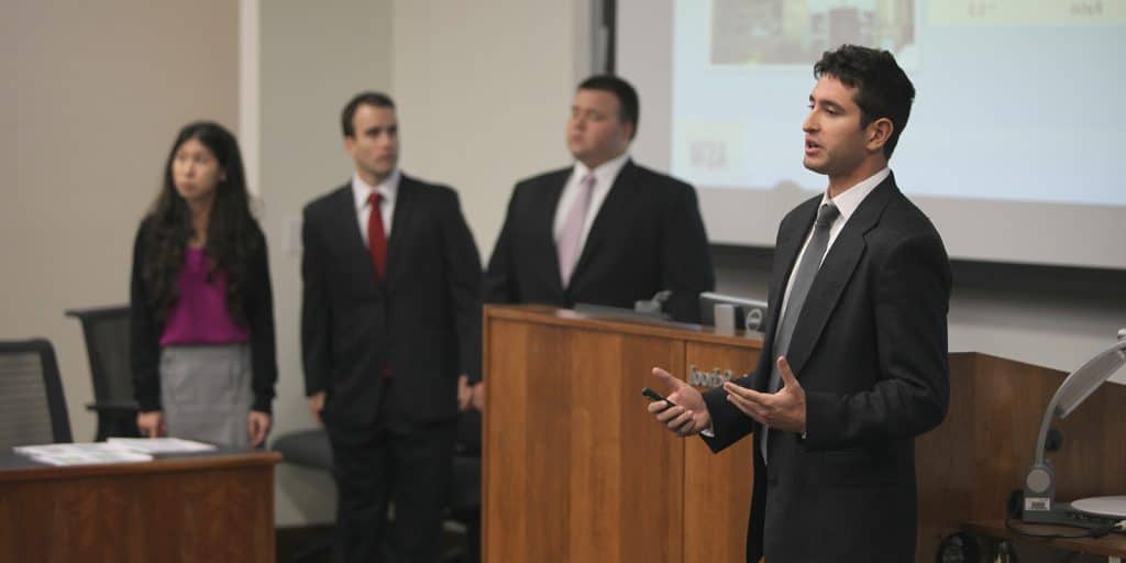 A group of older students in professional attire giving a presentation with the focus on a man towards the front.