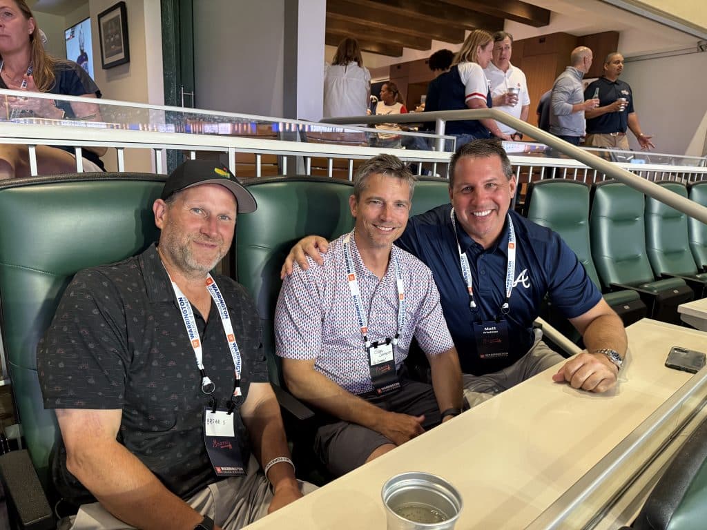 Three men wearing Atlanta Braves gear pose for a photo while watching a baseball game. 