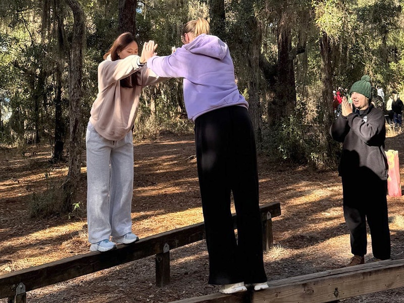 Two young women participate in a team building exercise with another woman to the side giving them directions.