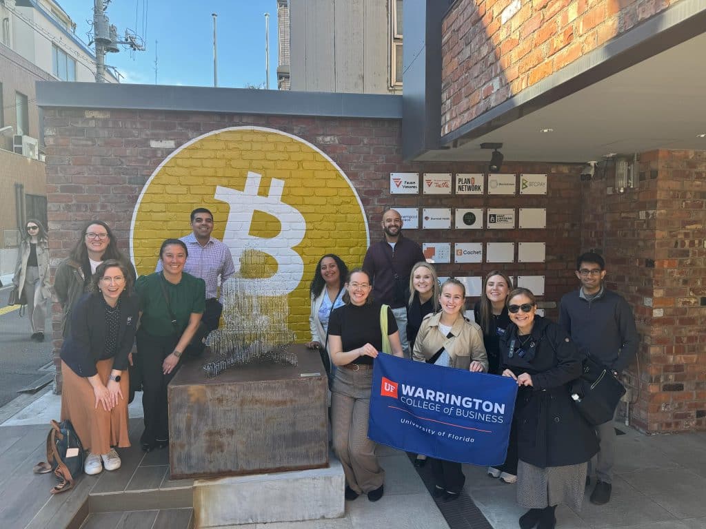 MBA student team in Tokyo standing and smiling in front of a yellow and white mural holding a Warrington banner