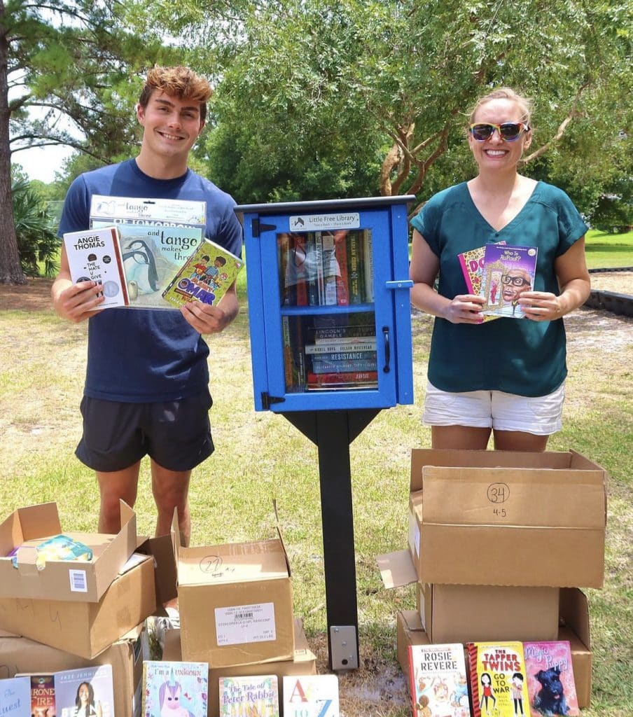 Chase Hartman poses with another person holding books next to a little library box stand.
