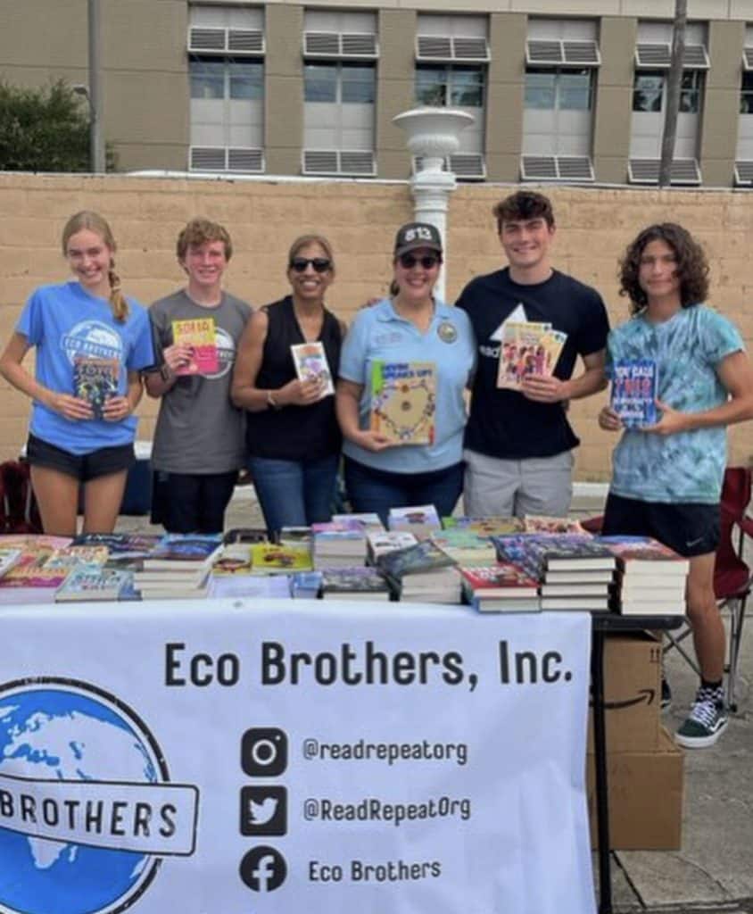 Chase Hartman stands with a group of people holding books in front of a table full of books with an Eco Brothers, Inc. banner.