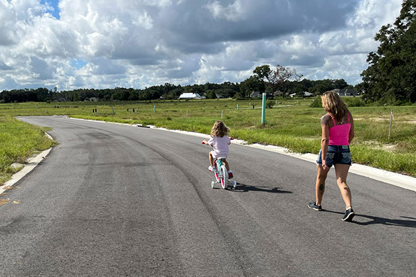A woman walks behind her daughter who is riding a small bike with training wheels along a quiet street where homes have not yet been built yet in Bridlewood