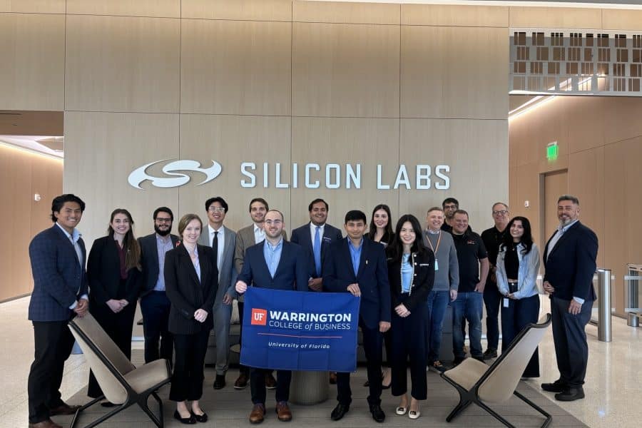 Students in Mike Carillo's MAN6930 Fall 2025 Pose in front of a Silicon Labs sign, holding a Warrington College of Business Flag.