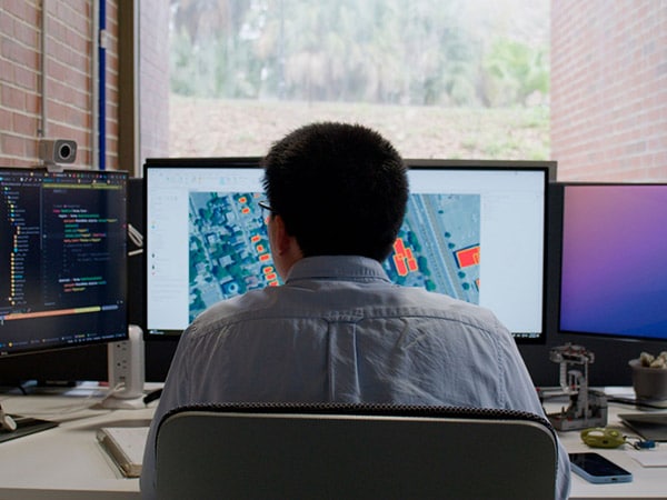 A researcher looks at his computer screen with an aerial image and structures highlighted in orange. Behind the computer screens is a window with brick walls on either side.