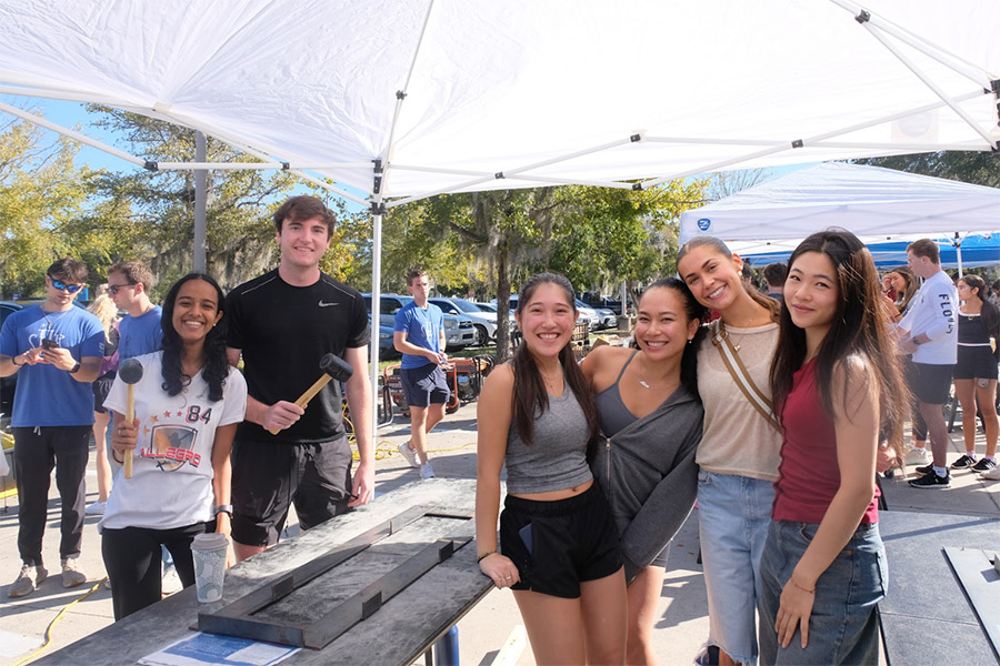 Six students smile under a both canopy on a sunny day as other students can be seen in the background.