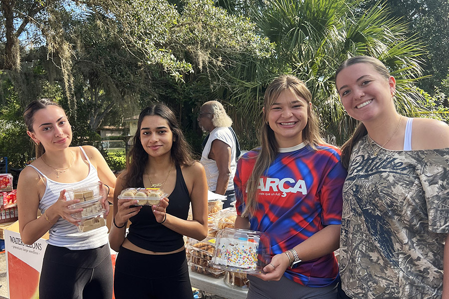 Four young women smile while three of them hold sweet desserts outside with a table behind them stacked with additional foot items.