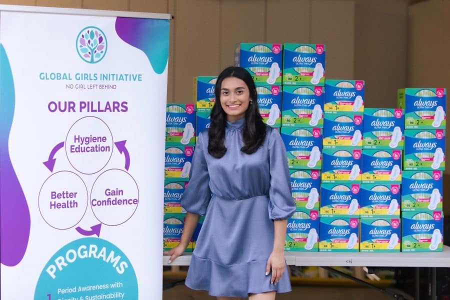 Aanya Patel poses in front of a box of donated menstrual products and a sign detailing the pillars of her nonprofit "Global Girls Initiative."