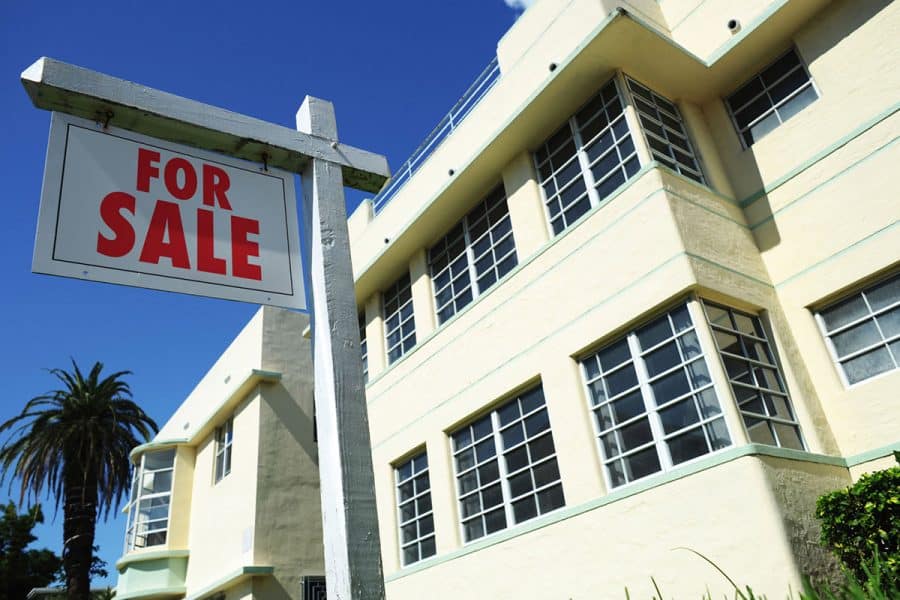 A pale yellow with light green trim art deco house in Miami with a for sale sign in front of it